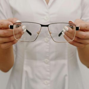 Crop anonymous female ophthalmologist in white medical robe showing stylish eyeglasses while working in light optometry clinic
